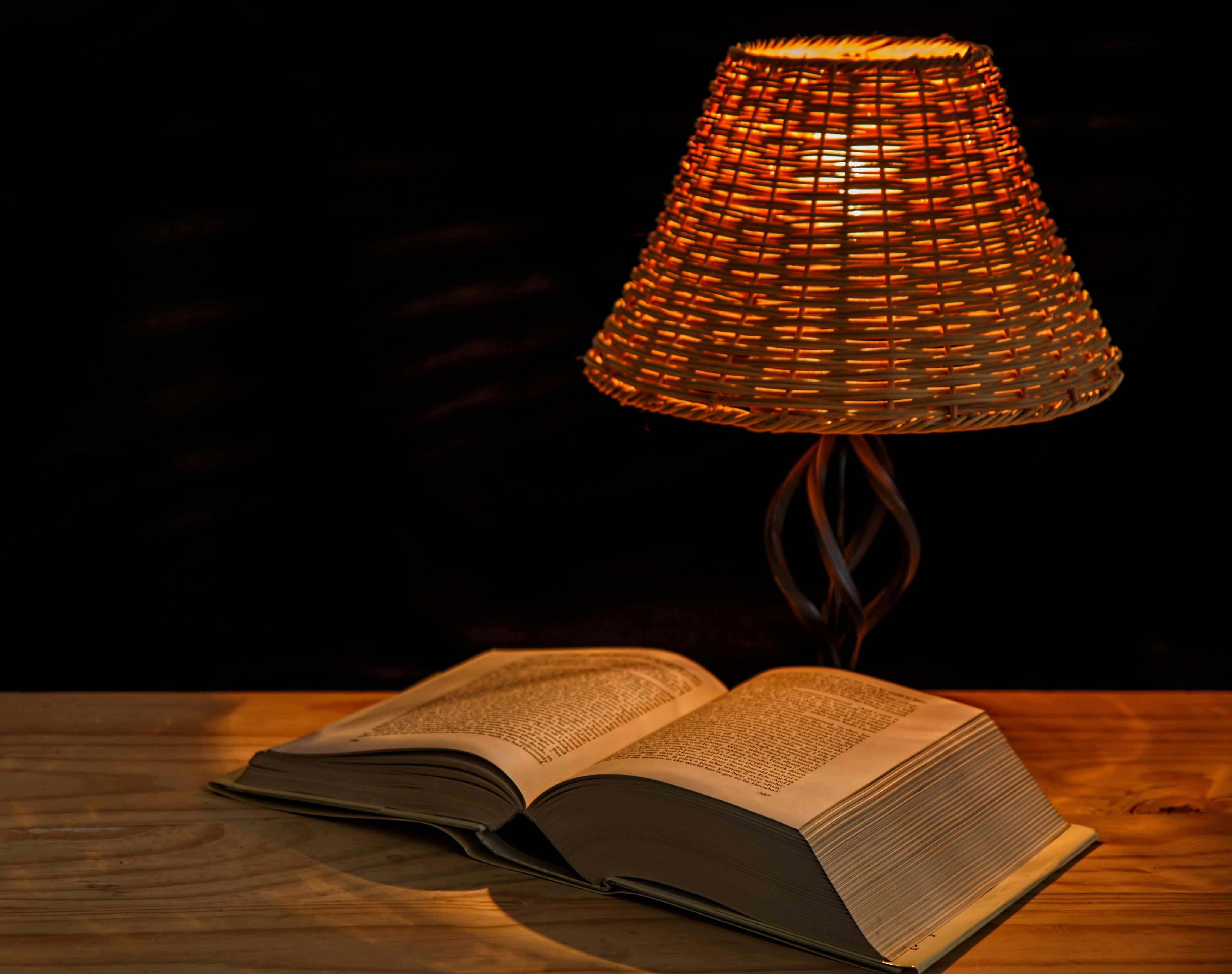 A linen-bound journal, a small ceramic lamp, and an open book on a wooden bedside table under warm evening light.